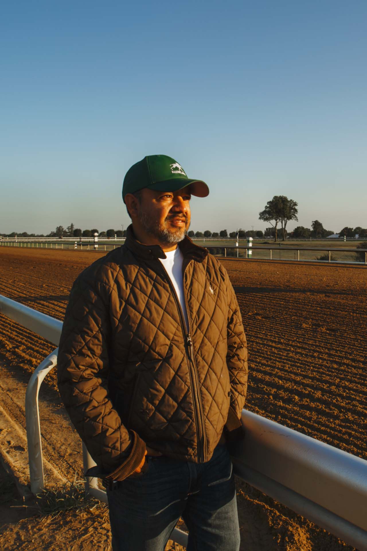 A photo of Alfredo Laureano looking off into the distance while standing by the dirt track at sunset. He is an older Hispanic man with graying chin stubble. He is wearing a green Elitehorses-brand hat and a brown Elitehorses-brand jacket overtop a white shirt.